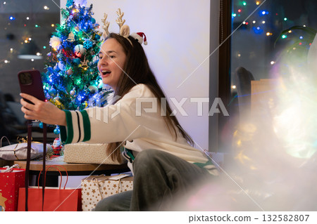Young woman with long hair making a video call near Christmas tree and gifts in cozy festive room Young woman with long hair making a video call near Christmas tree and gifts in cozy festive room 132582807