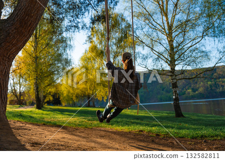 Woman swinging on wooden swing in spring park near lake under evening sunlight in Czech Woman swinging on wooden swing in spring park near lake under evening sunlight in Czech 132582811