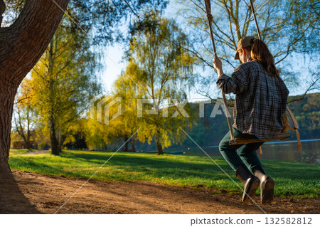 Woman swinging on wooden swing in spring park near lake under evening sunlight Woman swinging on wooden swing in spring park near lake under evening sunlight 132582812