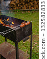 Grilled sausages on metal skewers over open fire in rustic outdoor grill with woodpile in background 132582833