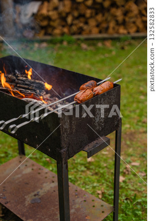 Grilled sausages on metal skewers over open fire in rustic outdoor grill with woodpile in background Grilled sausages on metal skewers over open fire in rustic outdoor grill with woodpile in background 132582833