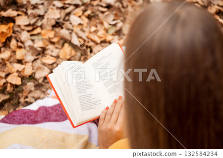 Reading in the park. Back shot of a girl reading some poems in colorful autumn park, sitting on the leaves and grass, wrapped in warm striped plaid 132584234