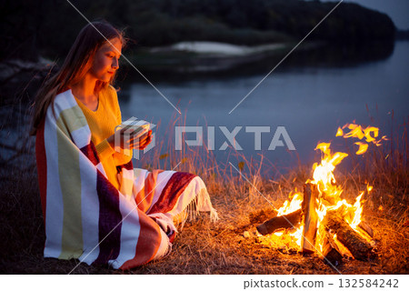 Young blond girl sitting near campfire on river bank and dreaming of something. Young girl drinking warm tea from her cup, admiring the calm evening near the river 132584242