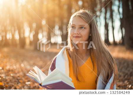 Beautiful girl reading a book and dreaming of something positive. Smiling blond girl likes to read books outdoors in autumn park 132584249