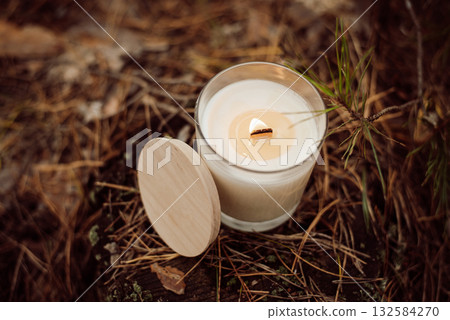 A glass with aroma candle with wooden wick on ground in spruce forest. Concept of aroma candle, pleasant smell of pine forest A glass with aroma candle with wooden wick on ground in spruce forest. Concept of aroma candle, pleasant smell of pine forest 132584270