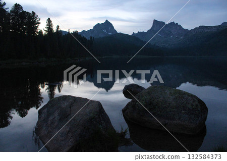 Silhouettes of two rocky mountain peaks and pine trees are reflected in the water of the Svetloye lake. Nature landscape in twilight in Ergaki nature park, Krasnoyarsk region, Siberia, Russia 132584373