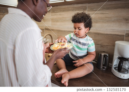Mother giving toddler healthy fruit snack in kitchen Mother giving toddler healthy fruit snack in kitchen 132585170
