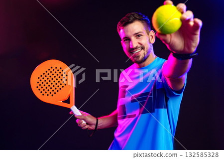 Young man plays padel tennis with racket and ball in colorful lighting Young man plays padel tennis with racket and ball in colorful lighting 132585328