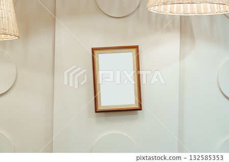 Empty wooden frame hanging on a white wall beneath woven light fixtures in a simple room 132585353