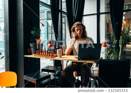 Young man working on laptop while talking on phone in a modern cafe setting 132585355