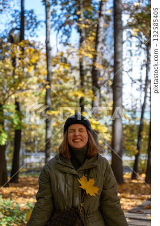 Woman in a green jacket and black beanie smiles joyfully in an autumn park with yellow leaves and sunlight filtering through the trees Woman in a green jacket and black beanie smiles joyfully in an autumn park with yellow leaves and sunlight filtering through the trees 132585405