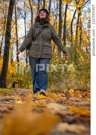 Woman walks through an autumn park in warm sunlight, wearing a khaki jacket and jeans surrounded by yellow leaves 132585407