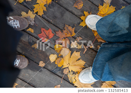 Close-up of two pairs of shoes standing on a wooden path covered with colorful autumn leaves in yellow, orange, and red tones Close-up of two pairs of shoes standing on a wooden path covered with colorful autumn leaves in yellow, orange, and red tones 132585413