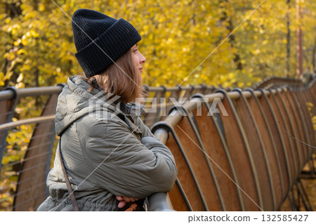 Woman in a gray jacket and black hat stands on a wooden bridge surrounded by golden autumn foliage, enjoying a peaceful moment in nature Woman in a gray jacket and black hat stands on a wooden bridge surrounded by golden autumn foliage, enjoying a peaceful moment in nature 132585427