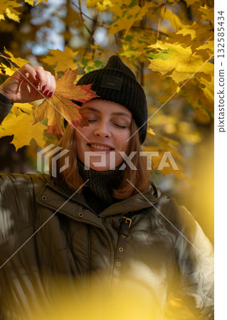 Woman in a green jacket and black beanie smiles with eyes closed, holding a yellow maple leaf over her face in an autumn park 132585434