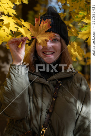 Smiling woman in a green jacket and black beanie playfully holds a yellow maple leaf over her eye in a sunlit autumn park Smiling woman in a green jacket and black beanie playfully holds a yellow maple leaf over her eye in a sunlit autumn park 132585435