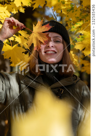 Smiling young woman in a green jacket and black beanie holds a yellow maple leaf over her eye in an autumn forest Smiling young woman in a green jacket and black beanie holds a yellow maple leaf over her eye in an autumn forest 132585436