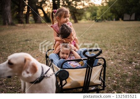Three sisters are playing in a wagon while their dog watches them Three sisters are playing in a wagon while their dog watches them 132585521