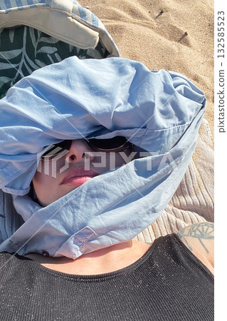 Young woman lying on beach sand with face wrapped in blue cloth and sunglasses, hiding from sun 132585523