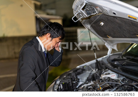 A man in a suit checks a car's malfunction on the road at night. He opens the hood of his car and looks helpless. 132586763
