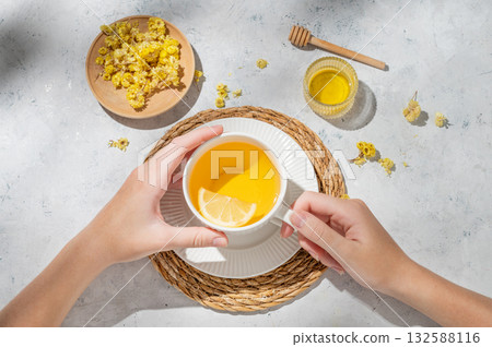 Hand holding cup of hot tea with lemon, honey and dry flowers on a white textured background  132588116
