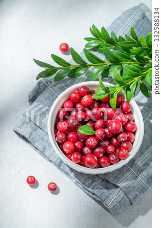 Fresh cranberries in a white bowl on a light background with green branch, napkin and sunlight. Fresh cranberries in a white bowl on a light background with green branch, napkin and sunlight. 132588134