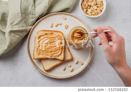 A hand holds a spoon with creamy peanut butter in a jar on a plate with toast and roasted nuts 132588202