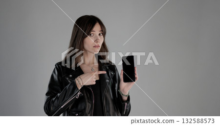 Young woman pointing at smartphone with blank screen on gray background Young woman pointing at smartphone with blank screen on gray background 132588573