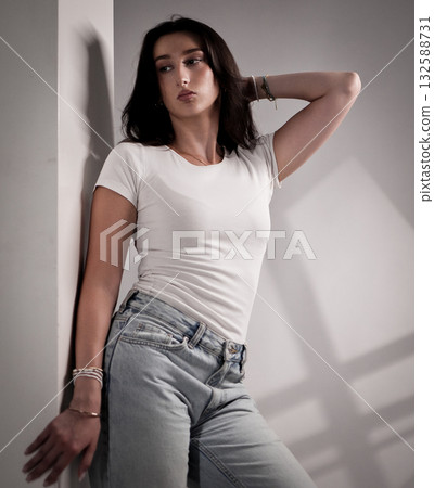 Young woman posing with hand on head, leaning against white wall, wearing jeans and white t shirt 132588731