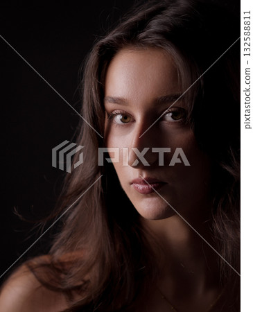 Portrait of young woman with long brown hair showing a serious expression against black background Portrait of young woman with long brown hair showing a serious expression against black background 132588811
