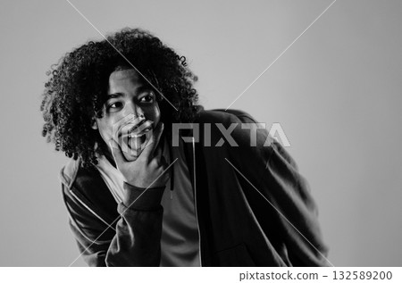 Young man with curly hair is making a funny face and sticking out his tongue while holding his chin 132589200