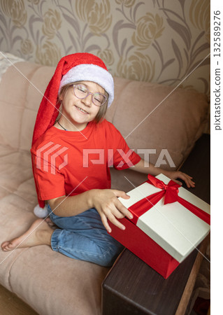 A girl in a Santa Claus hat opens a gift. It's Christmas time. A girl in a Santa Claus hat opens a gift. It's Christmas time. 132589276