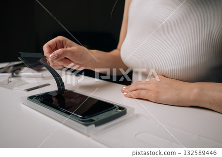 Cropped shot of woman installing screen protector on smartphone using specialized machine, ensuring perfect alignment and protection against scratches. Concept of mobile phone maintenance 132589446