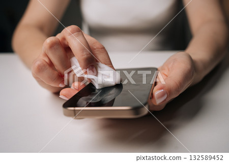 Close-up of unrecognizable woman cleaning smartphone screen with disinfectant wipe, ensuring hygiene and removing fingerprints and smudges from glass surface, closeup. Concept of healthy lifestyle. Close-up of unrecognizable woman cleaning smartphone screen with disinfectant wipe, ensuring hygiene and removing fingerprints and smudges from glass surface, closeup. Concept of healthy lifestyle. 132589452