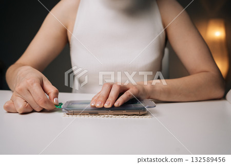 Close-up of woman applying screen protector to modern smartphone, ensuring precise alignment for optimal protection against scratches and potential damage to device display, sitting at white desk. Close-up of woman applying screen protector to modern smartphone, ensuring precise alignment for optimal protection against scratches and potential damage to device display, sitting at white desk. 132589456