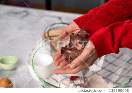 Close-up of hands holding a gingerbread man cookie cutter above a bowl of flour. Holiday baking preparation. Christmas cookie recipe, festive kitchen concept, home baking 132590008
