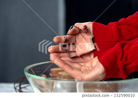 Close-up of hands holding a metal house-shaped cookie cutter. Holiday baking, cozy kitchen vibes, Christmas cookie preparation concept 132590010