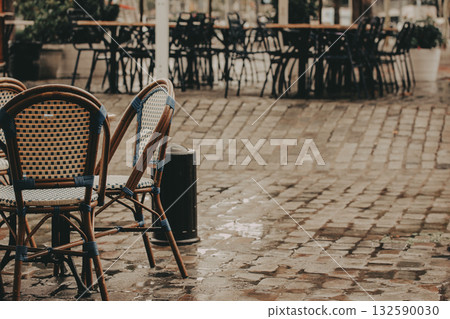 Rainy Day Cafe Scene. Wicker chairs and cobblestone streets create a cozy atmosphere in this outdoor cafe scene on a rainy day. Atmospheric city street scene with empty cafe terrace in a rainy day. 132590030