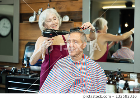 Elderly woman hairdresser dries hair with hairdryer to elderly male client. Elderly woman hairdresser dries hair with hairdryer to elderly male client. 132592316