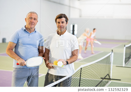 Portrait of two men pickleball players with rackets and ball standing on an indoor court Portrait of two men pickleball players with rackets and ball standing on an indoor court 132592329