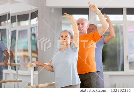 Happy smiling old woman dressed in sportswear enjoying ballet choreography, standing in fourth position at barre during group class in dance salon Happy smiling old woman dressed in sportswear enjoying ballet choreography, standing in fourth position at barre during group class in dance salon 132592355
