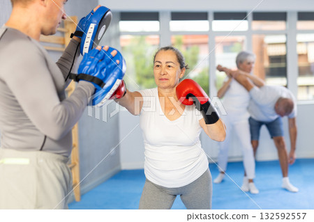 Senior lady and male couch are boxing in gym, man helps student to work out force of blow. 132592557