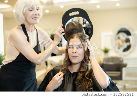 Older female hairstylist holding mirror presenting result of styling to young girl Older female hairstylist holding mirror presenting result of styling to young girl 132592581