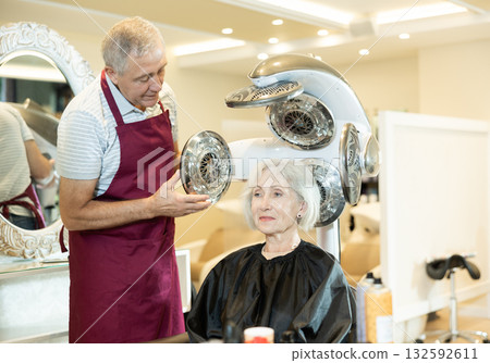 Experienced hairdresser adjusting settings of thermostimulator to dry hair for elderly woman 132592611
