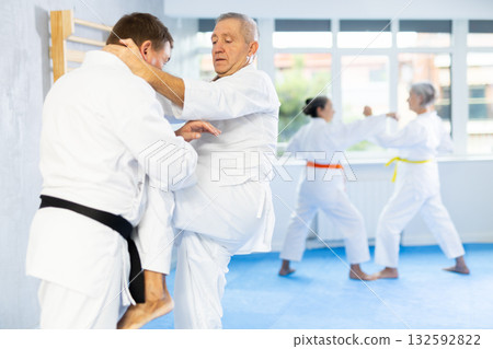 Old man attendee of karate classes fighting with his opponent in sports hall Old man attendee of karate classes fighting with his opponent in sports hall 132592822