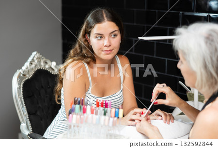 Friendly elderly woman working in beauty salon as manicure master shaping the nails of young female client with nail file Friendly elderly woman working in beauty salon as manicure master shaping the nails of young female client with nail file 132592844