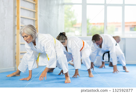 Group of mature women and men in kimonos doing muscle stretching before karate or judo class Group of mature women and men in kimonos doing muscle stretching before karate or judo class 132592877