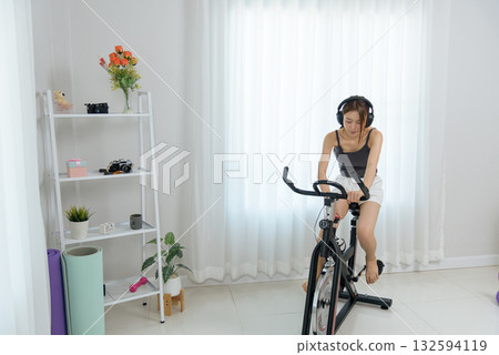 Young woman happy at home, people find wellbeing through activity, exercising on stationary bike with headphones, maintaining healthy lifestyle indoors Young woman happy at home, people find wellbeing through activity, exercising on stationary bike with headphones, maintaining healthy lifestyle indoors 132594119