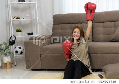 Young happy woman home, engages in activity people enjoy, celebrates sport victory with raised arm, wearing red boxing gloves, smiling after fitness training 132594146