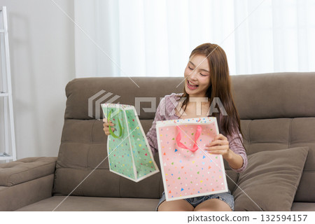 Young happy woman enjoying home activity for people, smiling, excited about gift bags, receiving presents, opening colorful shopping bags, sitting on comfortable sofa, experiencing joy and surprise 132594157